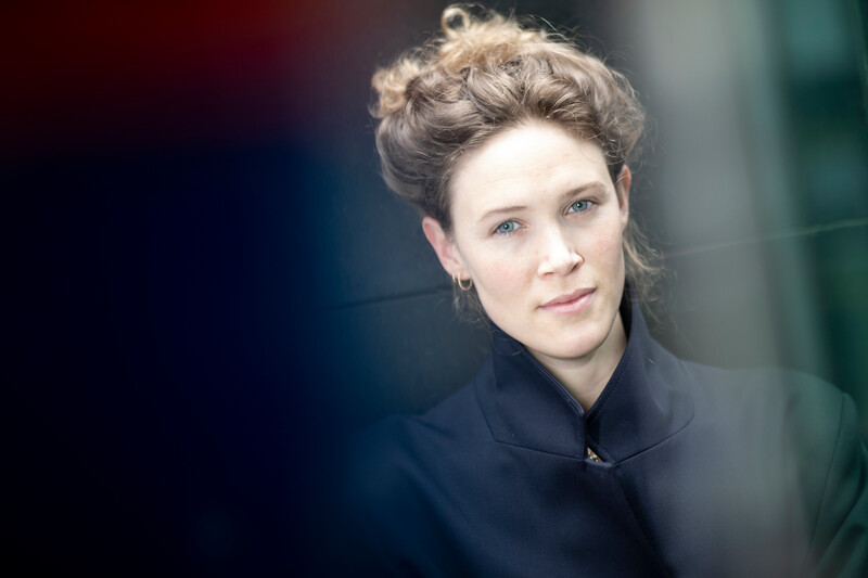 Dame med brunt hår i en høy dott, øynene smiler men hun ser seriøs ut, ser henne bare fra brystet og opp. Har på seg en svart kåpe som er kullet i halsen