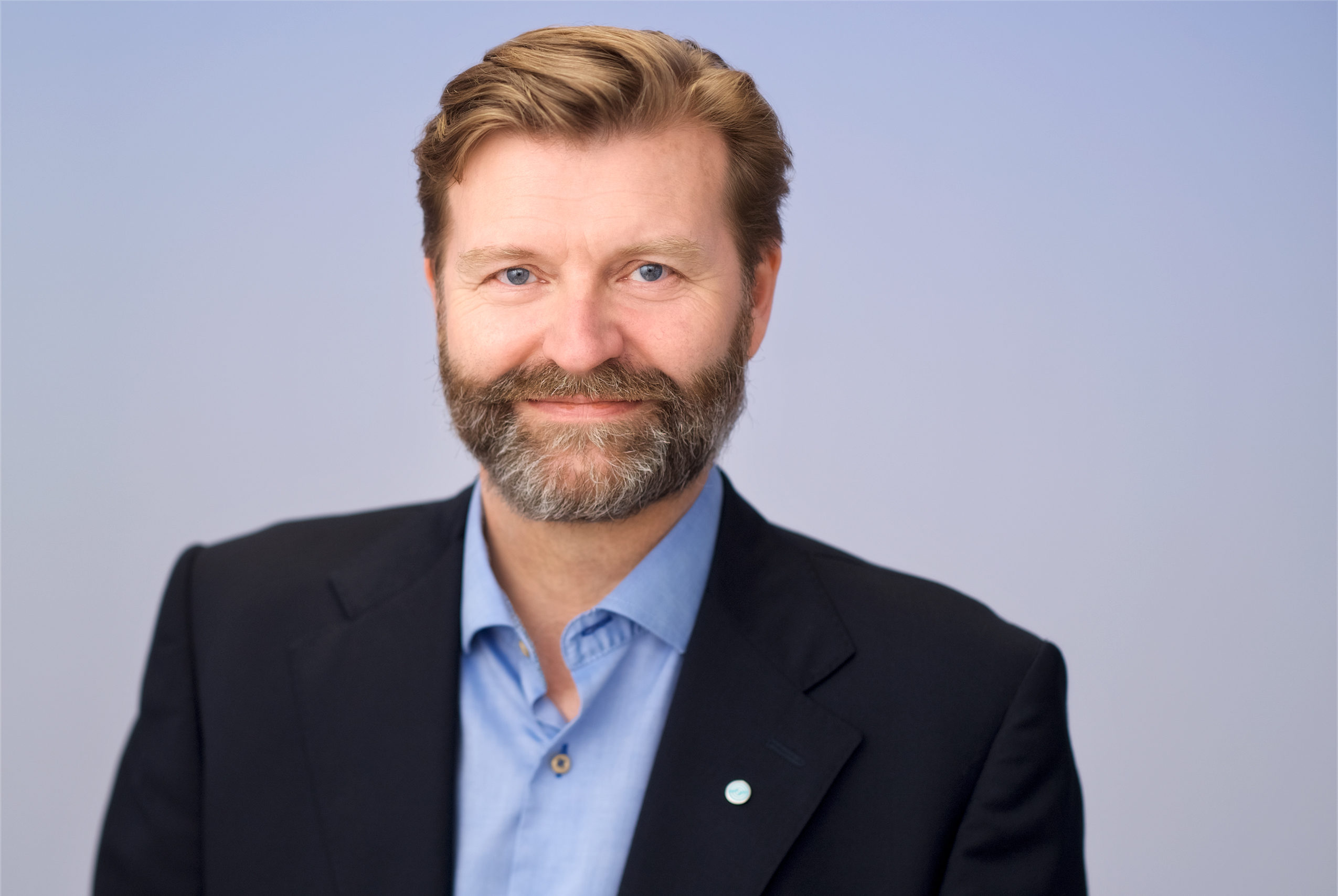 Mann med svart blazer blå skjorte, brunt hår og skjegg Blå bakgrunn
